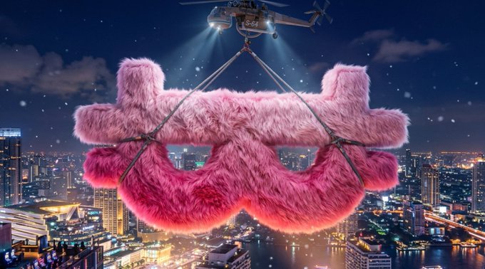 A large pink furry tripleS logo mask being carried by a helicopter over a city skyline at night.