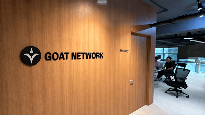 An office reception area with a wooden wall displaying the 'GOAT NETWORK' logo and a door labeled 'Mercury', with two people seated at desks in the background.