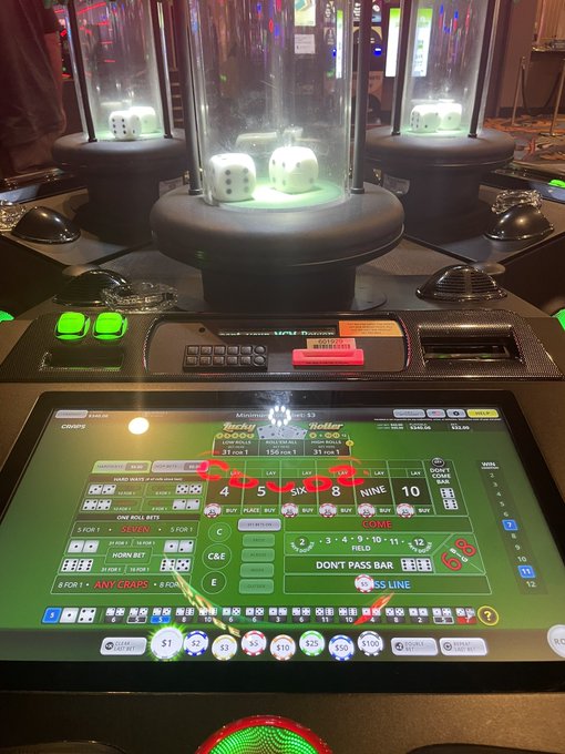 An electronic craps table game displays betting options including '4', '5 SIX 8', 'NINE', '10' on a green felt layout, with dice visible in an illuminated chamber above.