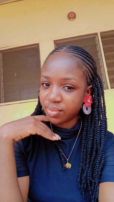 A young woman with long braided hair poses confidently in a navy blue t-shirt, accessorized with red flower earrings and an elephant pendant necklace, against a yellow building background—her poised, hopeful expression contrasts with the post's skeptical tone about who actually profits from NFTs.