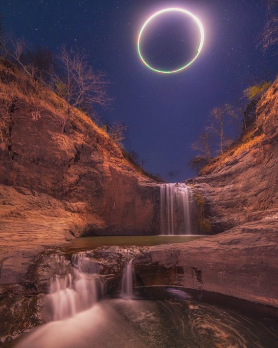 A composite astrophotography image featuring a glowing ring (possibly an eclipse or neon circle) in a starry night sky above a rocky waterfall scene.