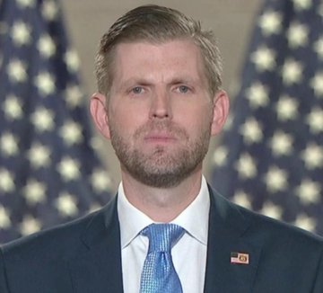 The image features a man wearing a suit, tie, and a pin with an American flag design. The man appears to be dressed for a formal event or occasion. The background of the image consists of a blue curtain adorned with white stars, which is likely part of a stage setting. The man's attire suggests that he might be attending a political event or ceremony related to President Trump and Bitcoin.