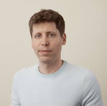 A photographic portrait shows a man with short reddish-brown hair and light eyes wearing a pale blue crew-neck sweater, posed against a plain off-white background with a subtle, neutral expression.