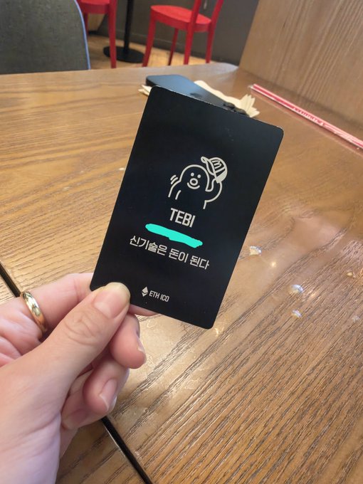 A hand holds up a black Ethereum (ETH ID) prepaid card with a cute bear mascot, displaying the name 'TEBI' and Korean text indicating the card has been activated.