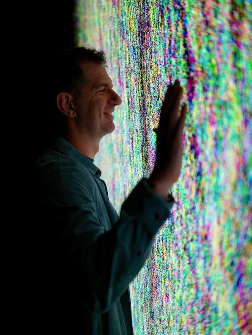 A man in a blue shirt smiles while reaching toward a vivid, pixelated rainbow light display, evoking themes of digital networks and interconnected nodes mentioned in the post about $TIBBIR. The vibrant cascade of colors behind him mirrors the colorful, web-like community being built by holders and users.