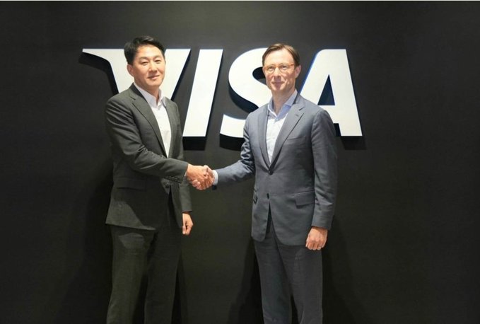 Two men in business suits shake hands in front of a large 'VISA' logo on a black background.
