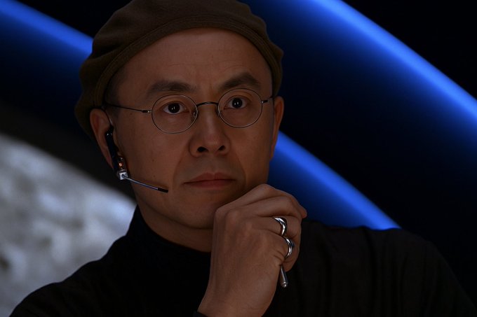 A close-up of an East Asian man wearing a dark beanie, round glasses, and a black turtleneck, with an earpiece microphone and rings on his finger, posed thoughtfully against a blue and gray background.