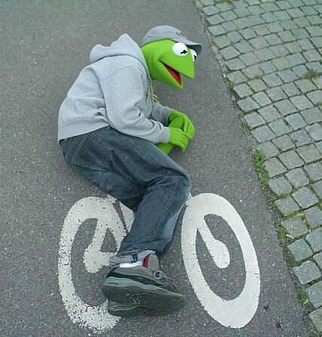 A person wearing a gray hoodie and jeans crouching on a street while wearing a Kermit the Frog mask, positioned over a painted bicycle lane symbol on cobblestone pavement.