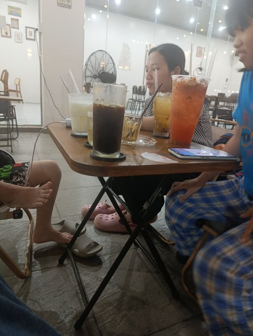 Three people sit around a small wooden table in a café, sharing four iced beverages with straws, including iced coffee and orange-colored drinks, with a smartphone resting on the table.