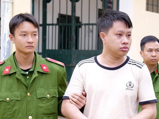 A man in a white 'Classic' t-shirt being escorted by police officers in green uniforms, appearing to be at a detention facility.