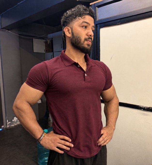 A muscular man with a beard and short curly hair wearing a maroon polo shirt, posing with his hands on his hips in what appears to be a gym setting.