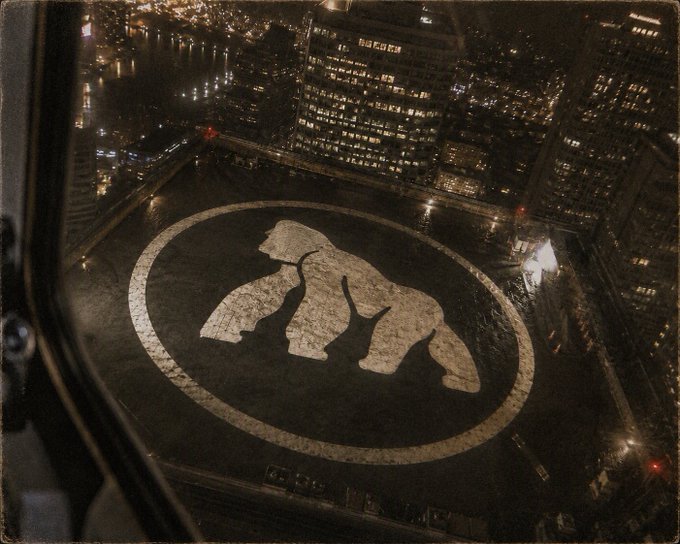 An aerial nighttime view shows a large circular emblem featuring three stylized apes displayed on a downtown plaza or rooftop. The illuminated three-ape logo reflects the iconic imagery of the Bored Ape Yacht Club, tying in with the "$APE" reference in the post as a playful nod to the cryptocurrency token associated with that NFT collection.