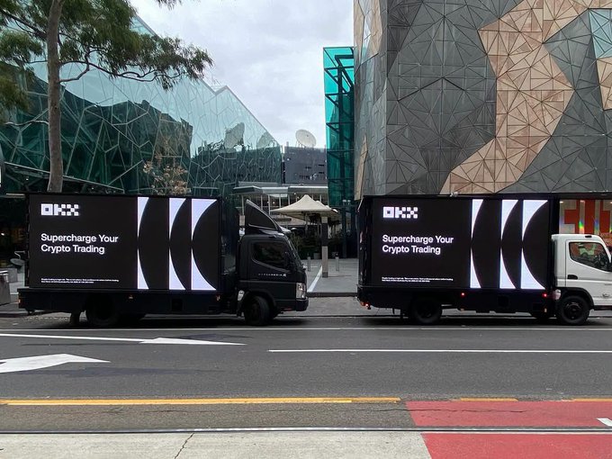 Two OKX trucks with mobile LED billboards reading "Supercharge Your Crypto Trading" are parked in front of modern geometric buildings, serving as mobile advertising for OKX's expansion into Europe with new licenses (MiCA, MiFID, PI) and an 8% deposit bonus up to €20,000 USDC for European users.