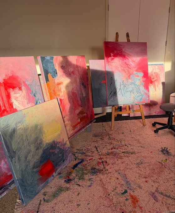 An artist's studio space filled with multiple abstract paintings in progress, featuring vibrant pinks, reds, blues, and yellows, displayed on easels and leaning against walls over a paint-stained drop cloth.
