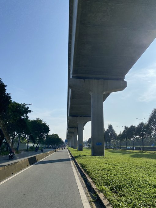A daytime view from underneath a massive concrete elevated highway or overpass, looking down a paved road with cyclists passing by on the left and green grass on the right.
