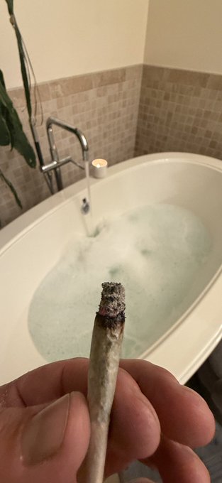 A hand holds a burnt joint in the foreground, with a white bathtub filled with bubbly water visible in the background.