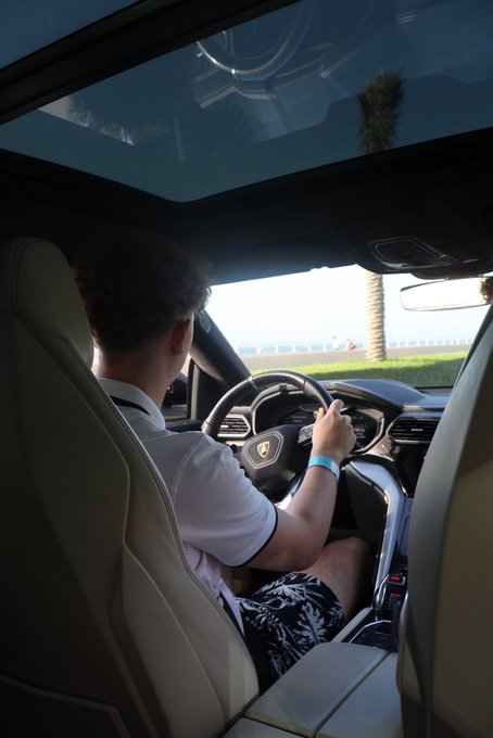 A young man drives a sleek sports car featuring a Lamborghini emblem on the steering wheel, fitting the "GM (get money)" lifestyle caption by showcasing a luxury vehicle through the windshield.