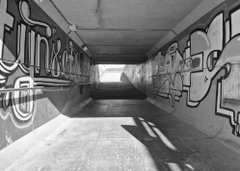 A black and white photograph shows the inside of a concrete tunnel covered in graffiti, leading toward a bright light at the end.
