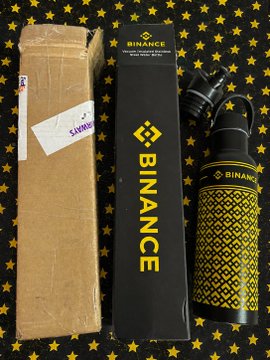 An unboxing photo showing a cardboard shipping box next to a black 'BINANCE' branded box and a matching black and yellow 'BINANCE' water bottle, displayed on a black surface with yellow stars.