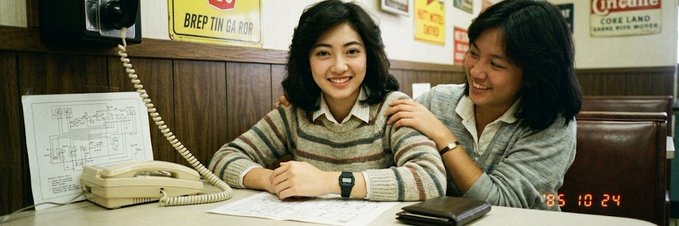 A vintage photograph dated '85 10 24 showing two smiling Asian women seated at a desk with a telephone, with signs in the background reading 'BREPTIN DA ROR' and 'Lucerne CORE LAND'.
