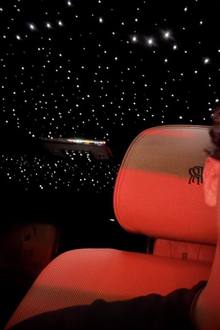A person sitting in a Rolls Royce vehicle (visible by the RR logo on the seat) at night, with a starry sky/ceiling display and what appears to be a model spaceship visible through the window.