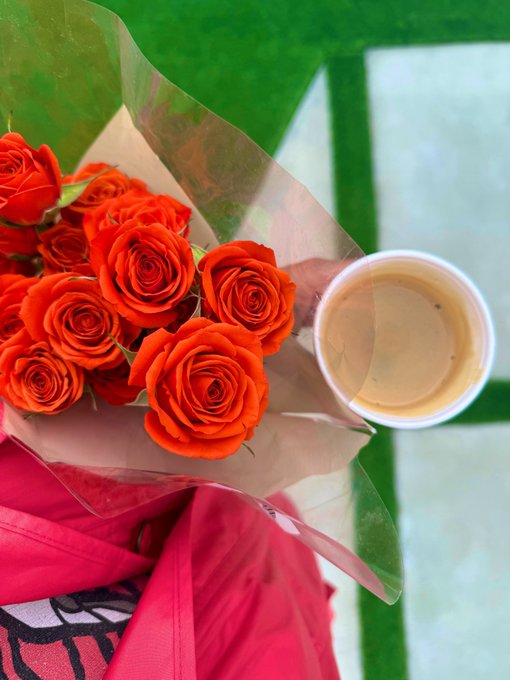 A bouquet of orange roses wrapped in brown paper with a pink ribbon, held next to a cup of milky coffee over a green and white surface.