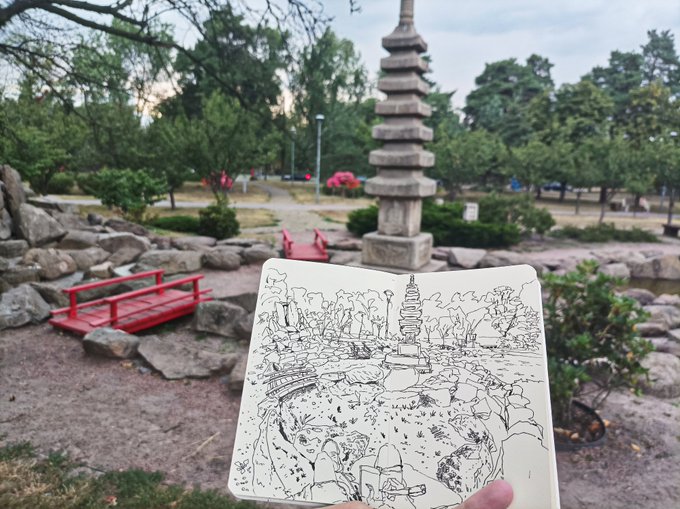 An open sketchbook in a park showing a detailed line drawing of a pagoda, red bridge, and rocky garden landscape that mirrors the real scene behind it, with a stone pagoda and red bridge visible in the actual park setting.
