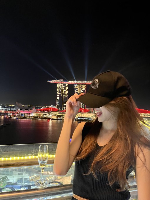 A woman in a black tank top and black cap with a silver crescent logo stands on a rooftop at night, holding a champagne glass with the illuminated Marina Bay Sands hotel and laser show in the background.