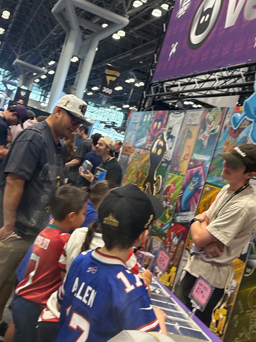 A photograph of a crowded convention floor, likely a gaming or trading card event, with people, including children in jerseys, gathered around a display of colorful artwork and merchandise.