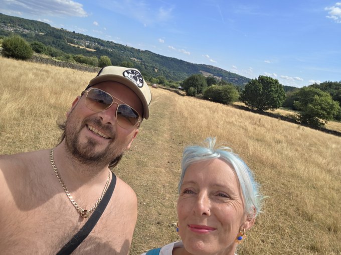 A selfie of a shirtless man wearing a "Bears" cap and sunglasses alongside a woman with light blue hair, standing in a grassy field with trees and hills in the background.