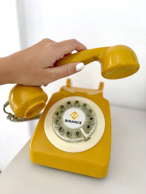 A hand lifts the receiver of a retro yellow rotary telephone marked with "BINANCE" on the dial center, visually reinforcing the post's "hello? giveaway is calling you…" message by playfully framing a giveaway as an incoming call.