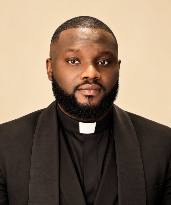 A professional portrait photograph of a Black man with a short beard wearing a black suit jacket over a clerical shirt with a white collar, suggesting he is a member of the clergy.