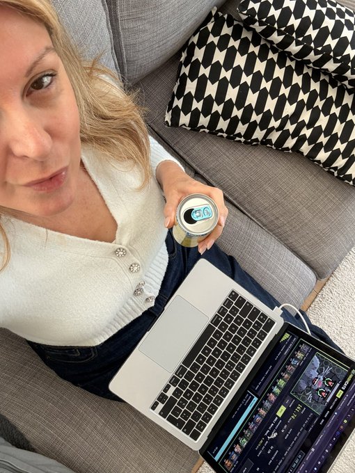A high-angle selfie of a blonde woman in a white cardigan sitting on a gray couch, holding a beverage can, with a laptop and an external monitor displaying video editing software (likely DaVinci Resolve) resting on her lap.