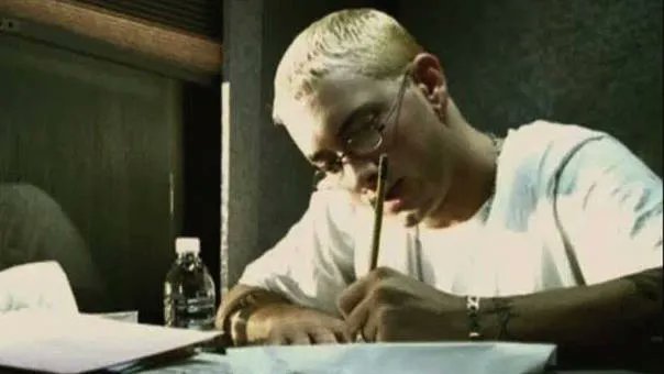A person with short blonde hair and glasses wearing a white shirt sits at a desk writing or drawing, with a water bottle nearby.