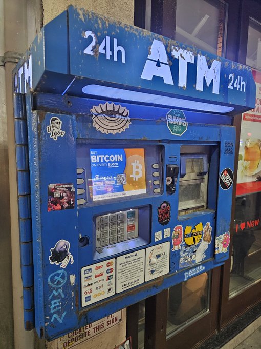 A blue 24-hour ATM machine, covered in various stickers and graffiti, advertises "BUY BITCOIN ON EVERY BLOCK" with a Bitcoin logo on its screen.
