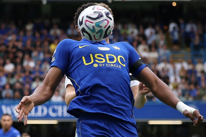 A Chelsea FC player heads a soccer ball during a Premier League match, with "USDC by Circle" prominently displayed on the front of his blue jersey. The image illustrates USDC's sponsorship visibility at one of the world's most-watched football leagues.