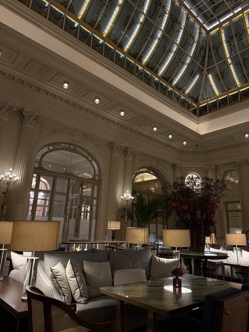 An elegant interior lounge features ornate ceilings, a glass skylight, plush sofas with pillows, and decorative lamps.