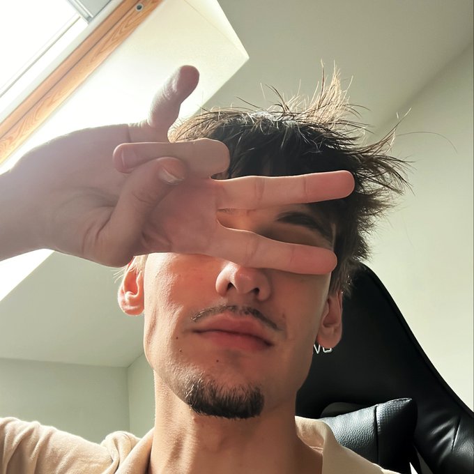 A young man with messy dark hair and a small goatee takes a close-up selfie, holding his hand up to partially cover his face in a casual pose. He appears to be relaxing indoors near a window, complementing the laid-back "Yo GANG 💎" greeting.