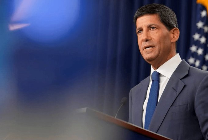 A man in a blue suit and tie standing at a podium, appearing to give a speech with a blue flag and American flag in the background.
