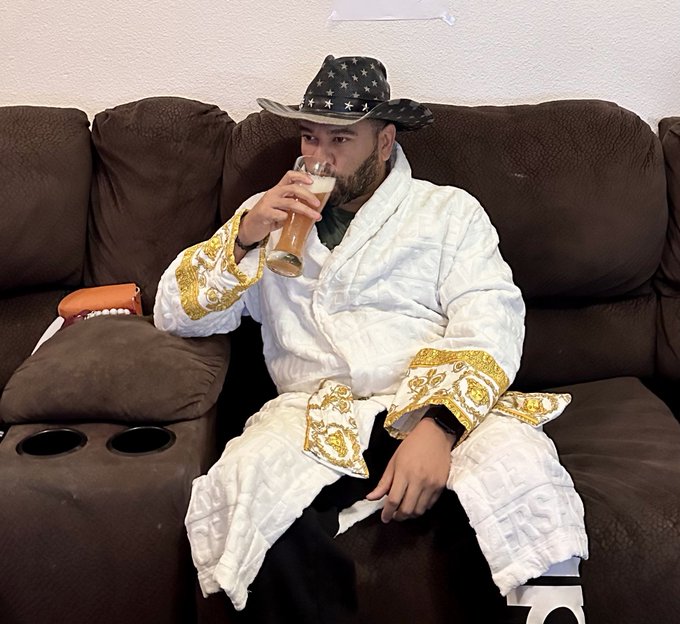 A bearded man wearing a Versace-style robe and an American flag cowboy hat sits on a brown couch drinking a beer.