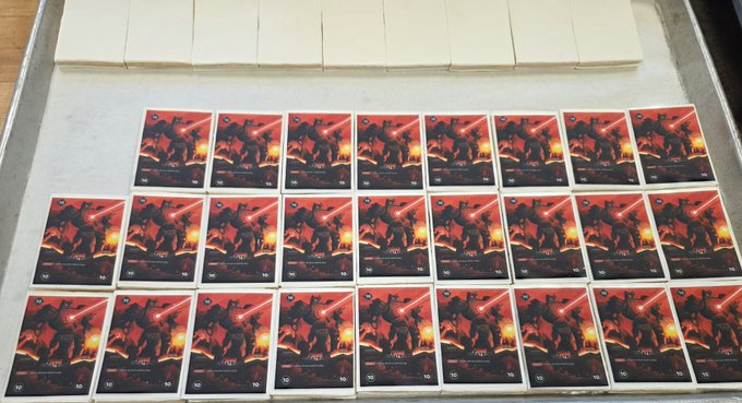 A neatly organized grid of approximately 24 identical "DOGINAL DOG LEGENDS" trading cards, each valued at "10" and featuring red-hued artwork of silhouetted warriors wielding lightsabers, laid out on a metallic surface beneath a row of blank white card backs. The repetitive presentation suggests a promotional or minting display for the DDLTCG.com card series mentioned in the post.