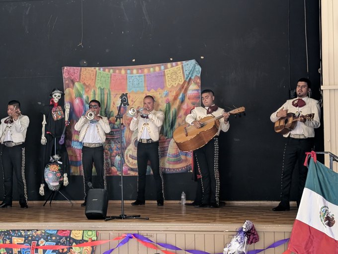 A mariachi band performs on stage with traditional attire, featuring trumpet, guitar, and vihuela players in front of a colorful Día de los Muertos tapestry backdrop, with a Mexican flag visible at the edge. The accompanying post mentioning "Xrp $850 in an hour" appears unrelated to the image content, suggesting a possible mismatch between the post and the celebratory cultural performance depicted.