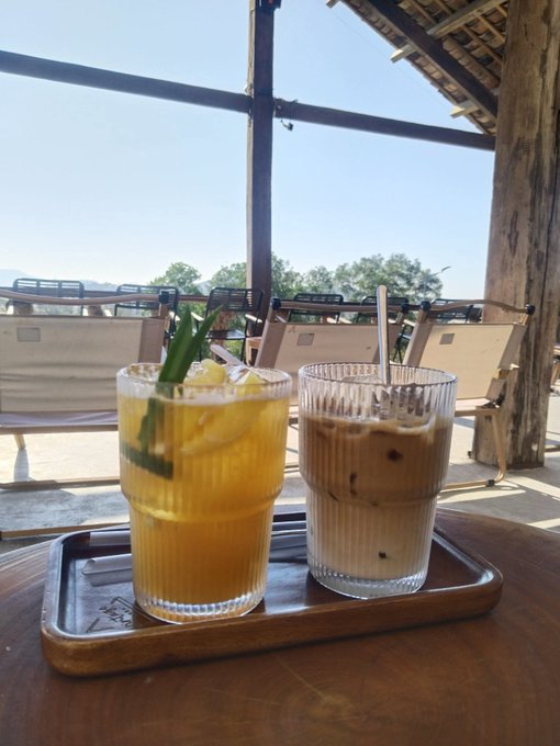 The image features two glasses of coffee sitting side by side on a wooden table, with one glass containing an iced coffee and the other filled with hot coffee. The background shows a view of trees through a window or a door, creating a serene atmosphere for enjoying their beverages.