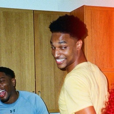 Two young men share a laugh in what appears to be a kitchen or indoor space, with one wearing a yellow shirt grinning broadly while the other in a blue shirt laughs heartily beside him. The "Farmercist rn" caption suggests a playful moment, possibly poking fun at their shared style or vibe as they react together.
