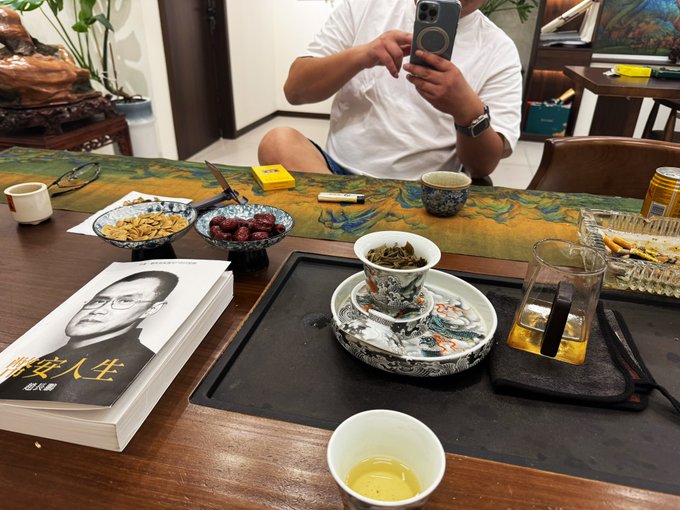 A person sitting at a wooden table taking a photo with a smartphone, with a traditional Chinese tea set, snacks (nuts and dates), and a book featuring a portrait of a man with Chinese text '平安人生' visible.