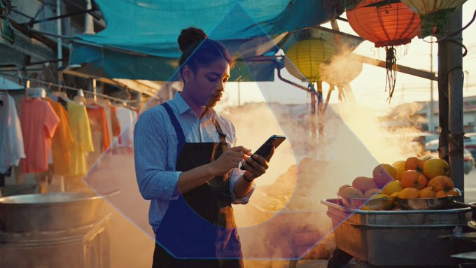A young woman in an apron uses her smartphone at a bustling market stall, surrounded by hanging clothes, fresh produce, and steaming pots, illustrating how everyday individuals can easily manage Bitcoin and stablecoin transactions on the go.