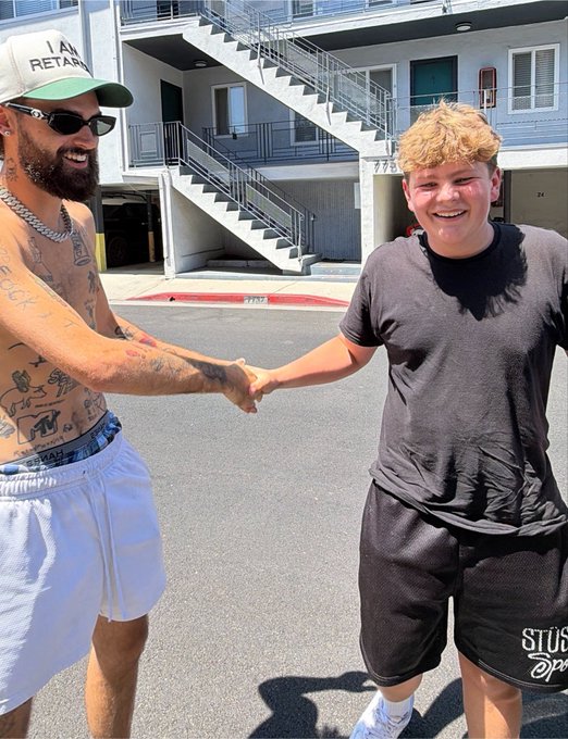 A shirtless tattooed man wearing an "I AM RETARDED" hat shakes hands with a smiling blonde kid in a black t-shirt outside an apartment building, with the watermark "STÜS... @pa..." visible on the shorts, referencing the "Stüssy trenches" meme kid lore mentioned in the post about him turning $20k into a legendary crypto cycle moment.