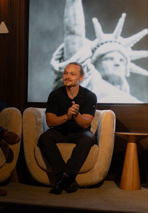 A man in a black polo shirt sits in a beige armchair with his hands clasped, posing in front of a large screen displaying the Statue of Liberty. The accompanying post notes he's updating his profile picture to this photo taken by @HodeyKo, apparently "borrowed" with appreciation.