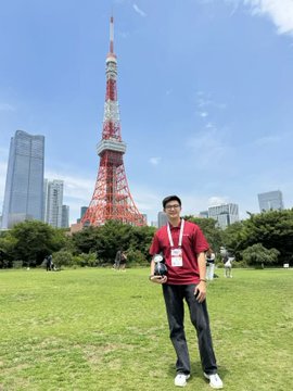 A man wearing a red shirt and lanyard posing on a grassy field with Tokyo Tower and city skyscrapers visible in the background on a sunny day.