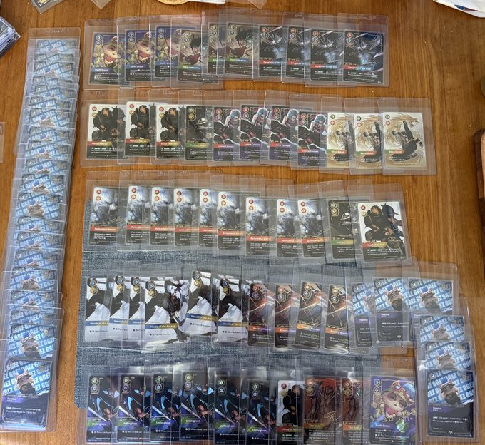 A collection of trading cards arranged in rows on a wooden floor, each individually sleeved in protective plastic covers, appearing to be from a strategy card game.